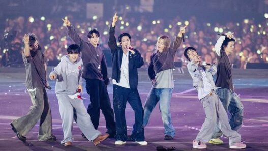 Enam anggota grup K-Pop ENHYPEN tampil energik di atas panggung konser besar dengan latar belakang penonton yang memegang lightstick menyala.