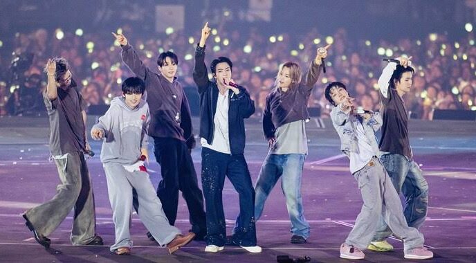 Enam anggota grup K-Pop ENHYPEN tampil energik di atas panggung konser besar dengan latar belakang penonton yang memegang lightstick menyala.