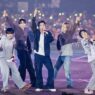 Enam anggota grup K-Pop ENHYPEN tampil energik di atas panggung konser besar dengan latar belakang penonton yang memegang lightstick menyala.