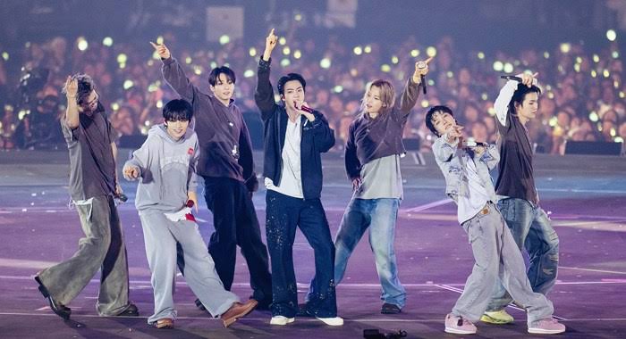 Enam anggota grup K-Pop ENHYPEN tampil energik di atas panggung konser besar dengan latar belakang penonton yang memegang lightstick menyala.