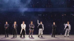 Tujuh anggota boy group K-Pop berpose penuh karisma mengenakan pakaian modis di atas panggung stadion megah berlatar lautan cahaya ungu.