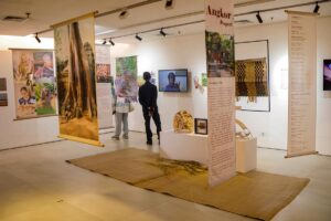 Pengunjung sedang melihat pameran seni bertema Angkor Project dengan spanduk gantung, instalasi kerajinan, dan layar video.