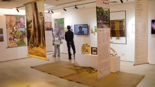Pengunjung sedang melihat pameran seni bertema Angkor Project dengan spanduk gantung, instalasi kerajinan, dan layar video.
