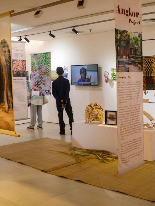 Pengunjung sedang melihat pameran seni bertema Angkor Project dengan spanduk gantung, instalasi kerajinan, dan layar video.