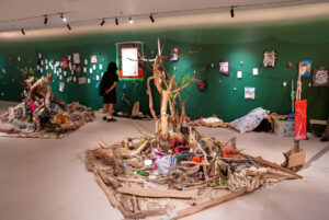 Ruang pameran seni kontemporer bernuansa dinding hijau yang menampilkan instalasi ranting kayu, sampah daur ulang, serta karya kertas yang tergantung.