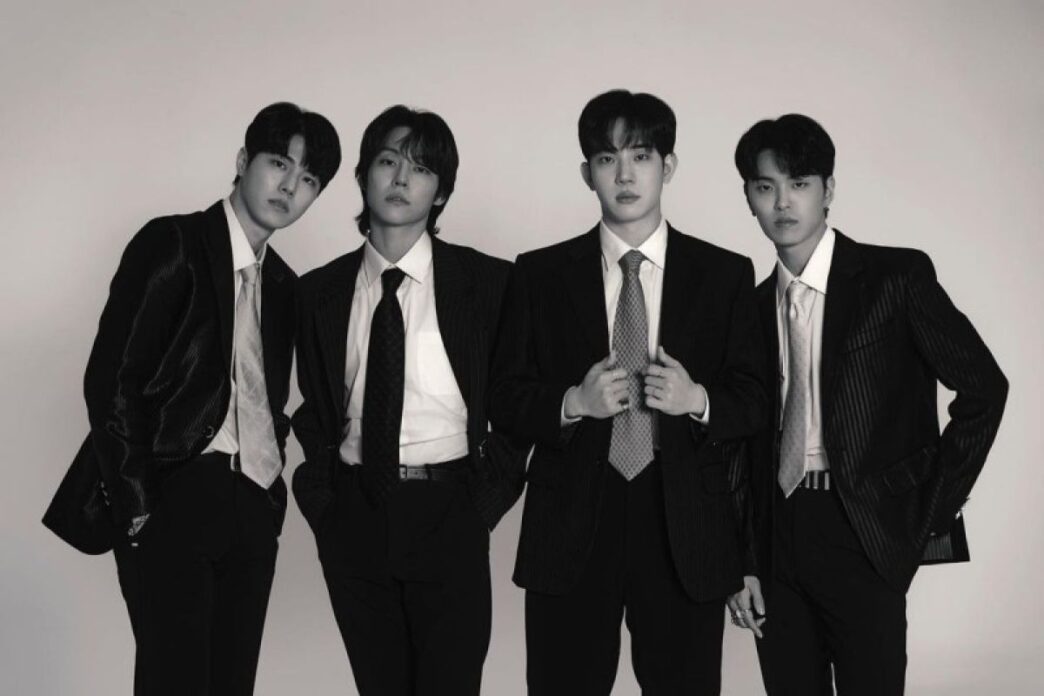 Foto hitam-putih empat anggota band DAY6 (Young K, Sungjin, Wonpil, dan Dowoon) berpose formal mengenakan setelan jas dan dasi rapi dengan latar belakang putih polos.