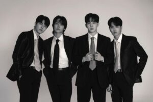 Foto hitam-putih empat anggota band DAY6 (Young K, Sungjin, Wonpil, dan Dowoon) berpose formal mengenakan setelan jas dan dasi rapi dengan latar belakang putih polos.