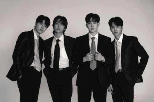 Foto hitam-putih empat anggota band DAY6 (Young K, Sungjin, Wonpil, dan Dowoon) berpose formal mengenakan setelan jas dan dasi rapi dengan latar belakang putih polos.