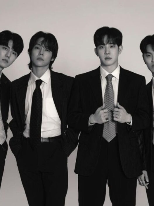Foto hitam-putih empat anggota band DAY6 (Young K, Sungjin, Wonpil, dan Dowoon) berpose formal mengenakan setelan jas dan dasi rapi dengan latar belakang putih polos.
