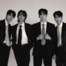 Foto hitam-putih empat anggota band DAY6 (Young K, Sungjin, Wonpil, dan Dowoon) berpose formal mengenakan setelan jas dan dasi rapi dengan latar belakang putih polos.