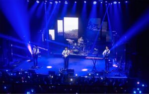 Penampilan panggung band The Rose di konser musik dengan pencahayaan spektakuler berwarna biru dramatis, menampilkan vokalis, gitaris, bassist, dan drummer di hadapan penonton.