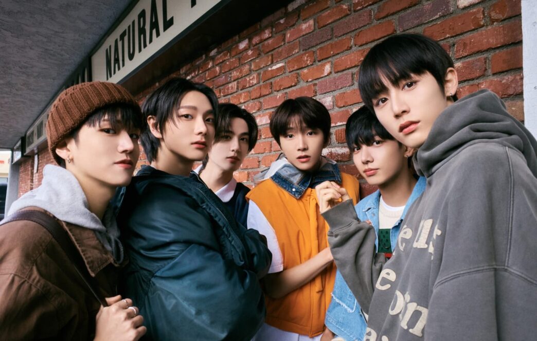 Enam anggota boyband K-pop BOYNEXTDOOR berpose gaya casual streetwear di luar ruangan dengan latar belakang dinding bata merah.