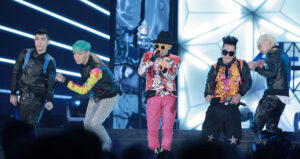 Lima anggota grup K-pop BIGBANG (Seungri, T.O.P, G-Dragon, Taeyang, dan Daesung) beraksi energik di atas panggung konser dengan pakaian colorful bergaya hip-hop eksentrik.