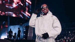 Ye (Kanye West) tersenyum mengenakan jaket putih, kacamata hitam, dan sarung tangan hitam di atas panggung konser