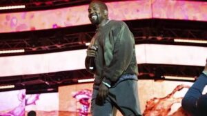 Musisi Ye (Kanye West) memegang mikrofon dan tersenyum di atas panggung berlatar layar LED berwarna merah muda