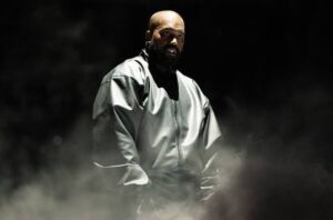 Musisi Ye (Kanye West) mengenakan jaket perak metalik berdiri di panggung konser berlatar gelap dan asap imersif