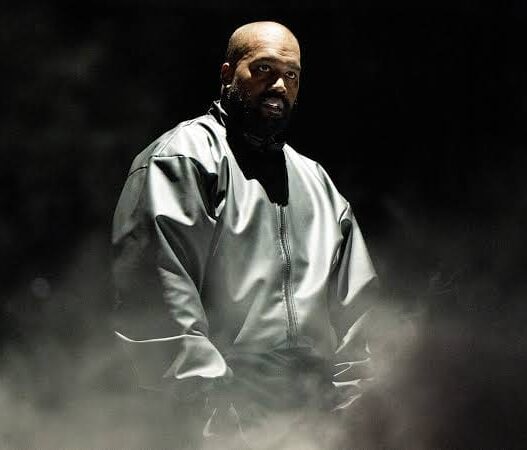 Musisi Ye (Kanye West) mengenakan jaket perak metalik berdiri di panggung konser berlatar gelap dan asap imersif