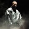 Musisi Ye (Kanye West) mengenakan jaket perak metalik berdiri di panggung konser berlatar gelap dan asap imersif