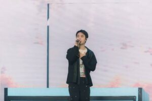Doh Kyung Soo (D.O. EXO) bernyanyi di atas panggung dengan memegang mikrofon di tangan kanan dan meletakkan tangan kirinya di dada. Ia mengenakan luaran hitam, kaus bermotif, dan celana hitam beraksen bintik-bintik cerah dengan latar belakang panggung bernuansa terang.
