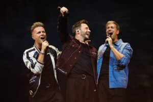 Tiga personel grup musik Westlife (Nicky Byrne, Shane Filan, dan Kian Egan) bernyanyi bersama di atas panggung konser sambil memegang mikrofon dengan ekspresi gembira.