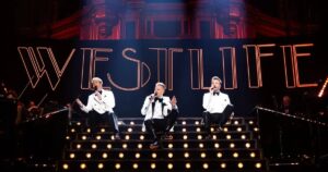 Tiga anggota grup musik Westlife mengenakan setelan jas tuksedo putih dan dasi kupu-kupu hitam duduk bernyanyi di tangga panggung berlampu dengan tulisan neon besar WESTLIFE di latar belakang.