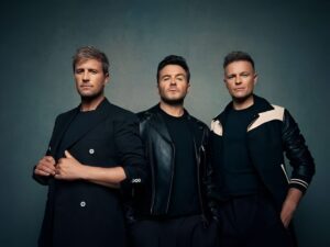 Tiga anggota grup musik Westlife (Kian Egan, Shane Filan, dan Nicky Byrne) berdiri bersama dalam pakaian hitam berpose untuk foto studio.
