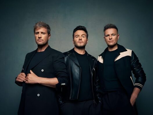 Tiga anggota grup musik Westlife (Kian Egan, Shane Filan, dan Nicky Byrne) berdiri bersama dalam pakaian hitam berpose untuk foto studio.