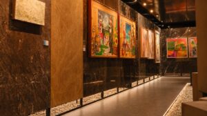 Lorong galeri seni bernuansa hangat dan elegan dengan dinding batu alam gelap serta pencahayaan sorot yang menampilkan karya lukisan kontemporer berbingkai kayu.