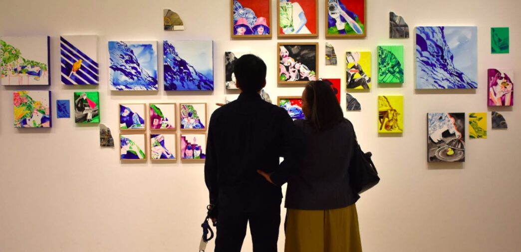 Dua orang pengunjung tampak dari belakang sedang berdiri dan mengamati dinding pameran yang dipenuhi puluhan lukisan kontemporer berwarna-warni berukuran kecil hingga sedang.