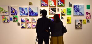 Dua orang pengunjung tampak dari belakang sedang berdiri dan mengamati dinding pameran yang dipenuhi puluhan lukisan kontemporer berwarna-warni berukuran kecil hingga sedang.
