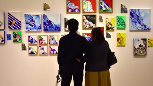 Dua orang pengunjung tampak dari belakang sedang berdiri dan mengamati dinding pameran yang dipenuhi puluhan lukisan kontemporer berwarna-warni berukuran kecil hingga sedang.