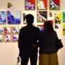 Dua orang pengunjung tampak dari belakang sedang berdiri dan mengamati dinding pameran yang dipenuhi puluhan lukisan kontemporer berwarna-warni berukuran kecil hingga sedang.