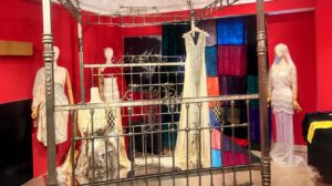 Instalasi pameran busana dan seni di dalam ruang berdinding merah, menampilkan gaun-gaun kontemporer anggun yang dipajang pada manekin serta struktur ranjang besi berhias di bagian depan.