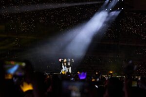 Sekelompok penampil berdiri di atas panggung di bawah sorotan lampu sorot dalam konser dengan lautan penonton yang memegang lightstick.