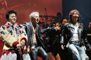 Beberapa musisi pria tampil di atas panggung konser dengan pakaian penuh gaya dan ekspresi ceria.